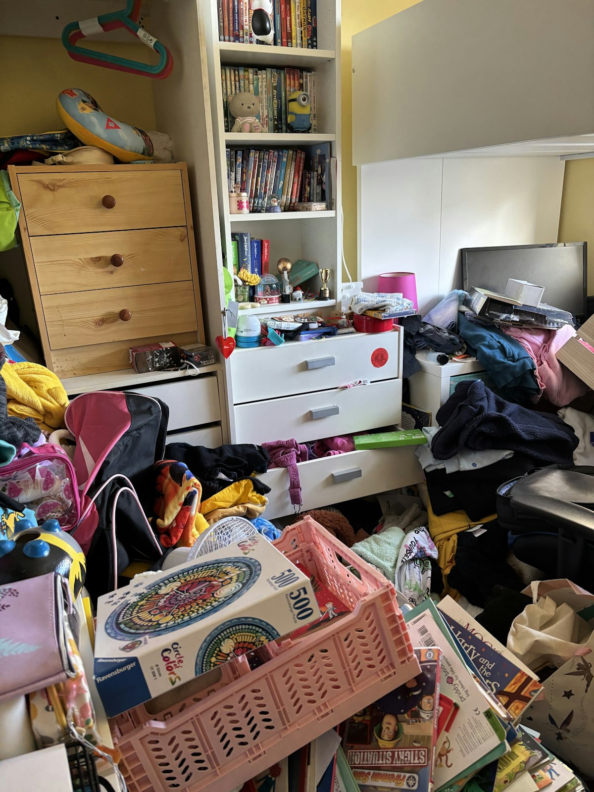 Cluttered child's bedroom with toys, clothes and belongings scattered across floor and furniture
